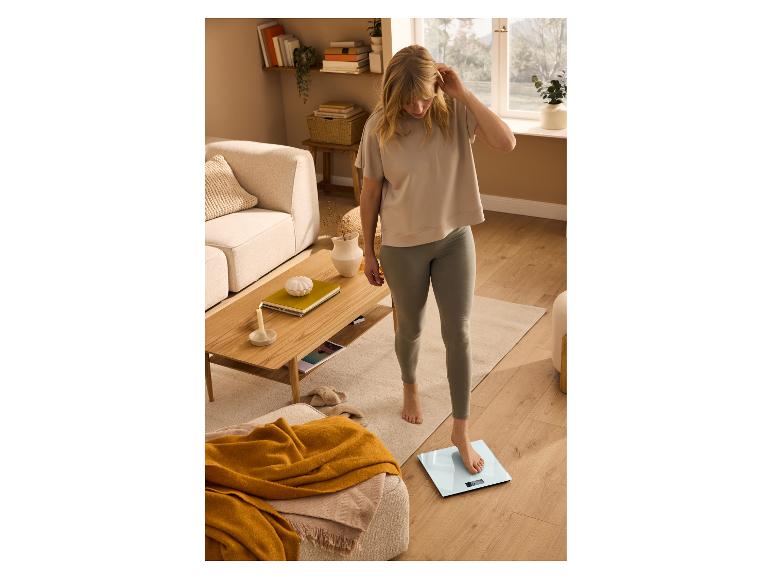 Jeune femme en t-shirt et leggings, se pesant sur une balance de salle de bain numérique.