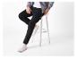 Homme en pantalon cargo noir, t-shirt blanc et veste zippée grise, assis sur un tabouret blanc.