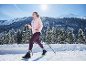 Femme en veste rose et leggings violets faisant de la marche nordique dans un paysage enneigé.