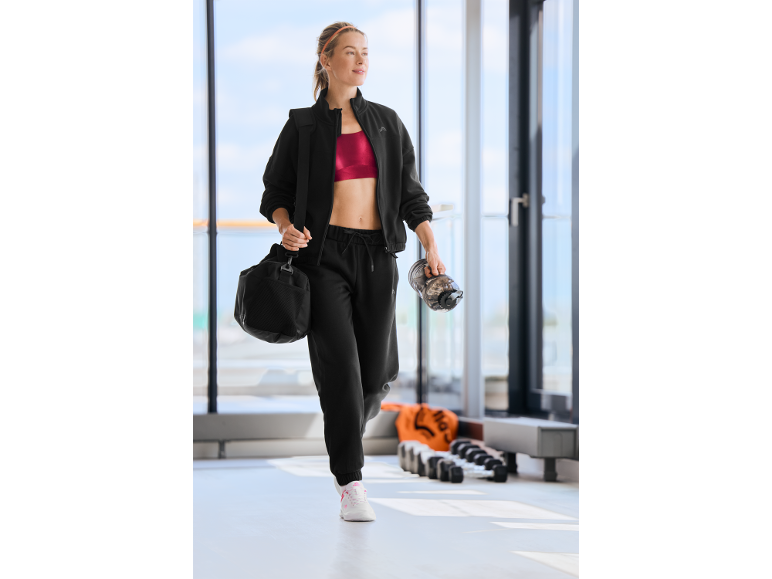 Une femme en survêtement noir et brassière de sport rouge marche dans une salle de sport.