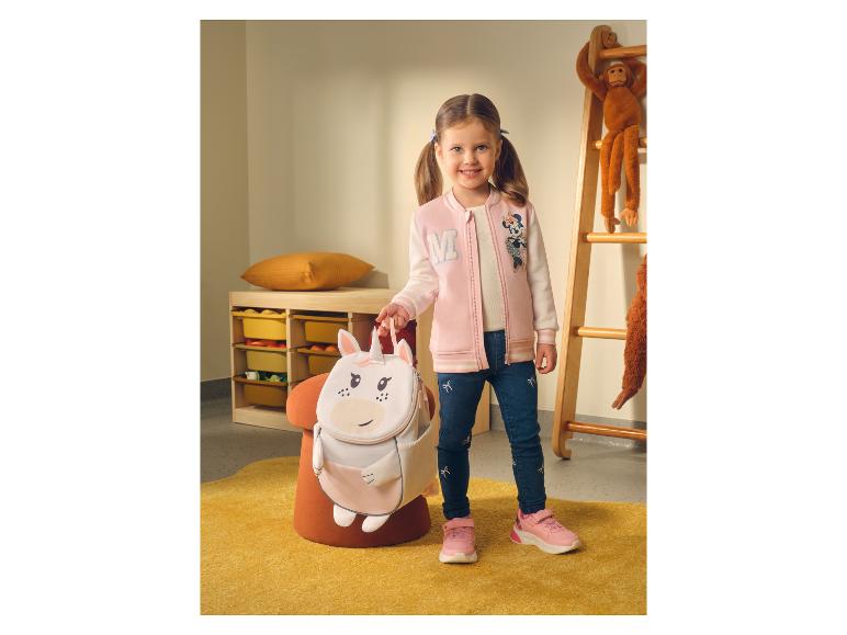 Fillette souriante en veste rose et jean, tenant un sac à dos licorne.