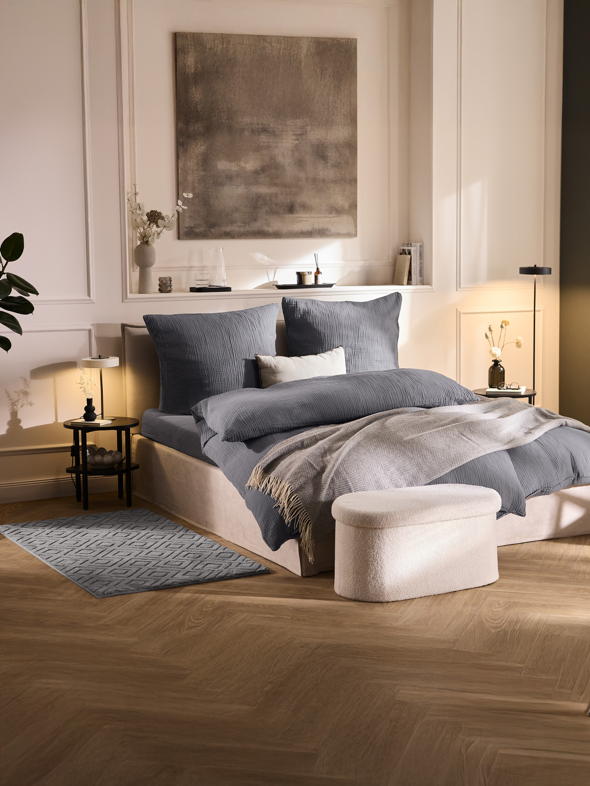 Chambre avec un lit doté de linge de lit gris, d'un jeté gris clair et d'un pouf crème.