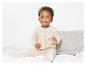 Un jeune enfant vêtu d'un cardigan et d'un pantalon en tricot beige est assis sur un lit blanc.