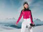 Femme portant un pull rouge et noir et un pantalon de ski blanc Craft, debout devant des montagnes.