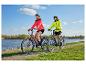 Couple à vélo électrique le long d'une rivière, portant des vestes de sport et des casques.
