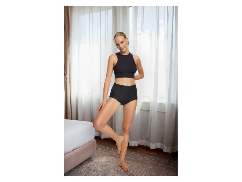 Femme en sous-vêtements noirs, haut court et culotte taille haute, posant dans une chambre.