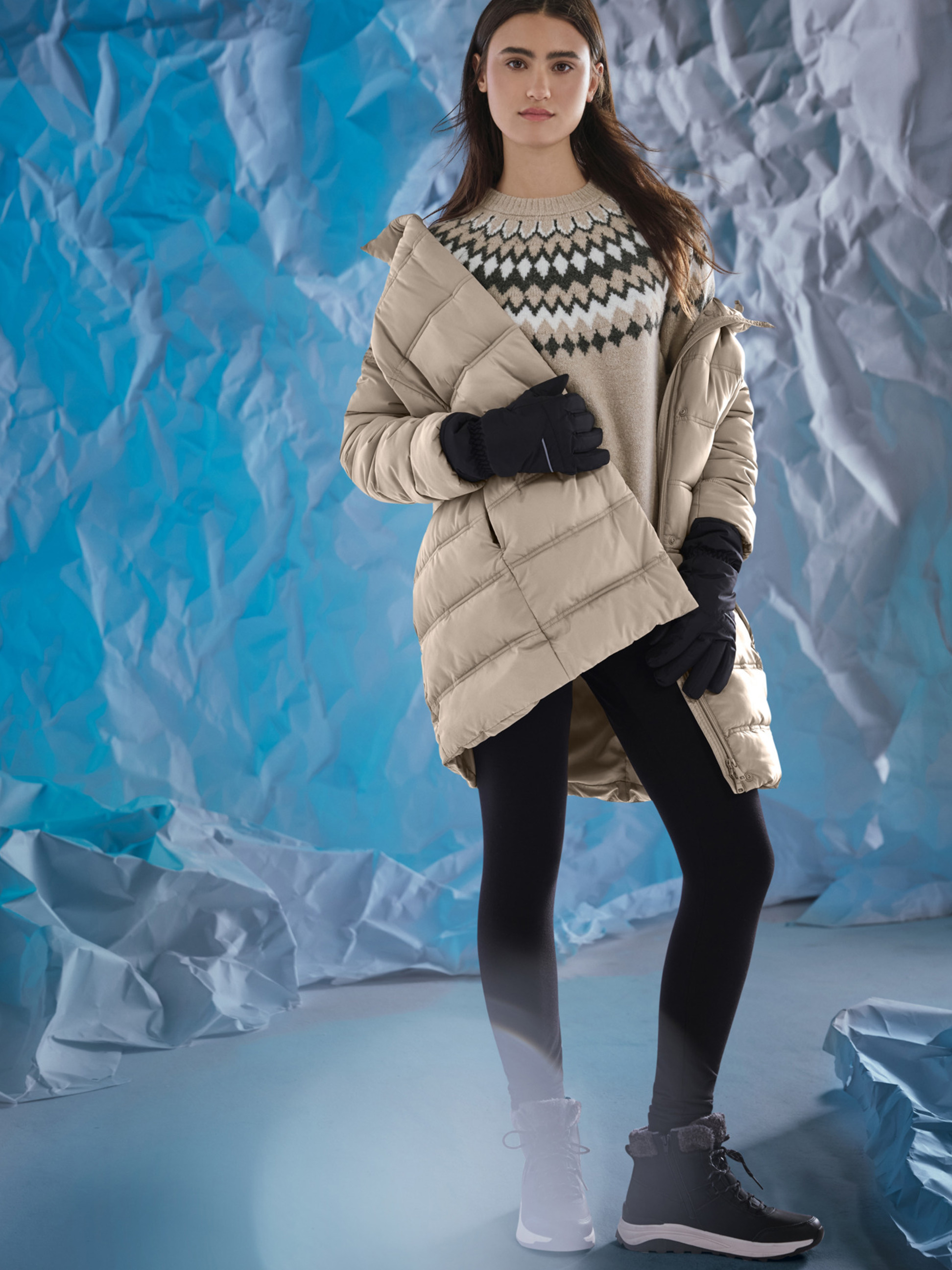 Femme en doudoune beige, pull à motifs, leggings noirs et bottes d'hiver, sur fond bleu.