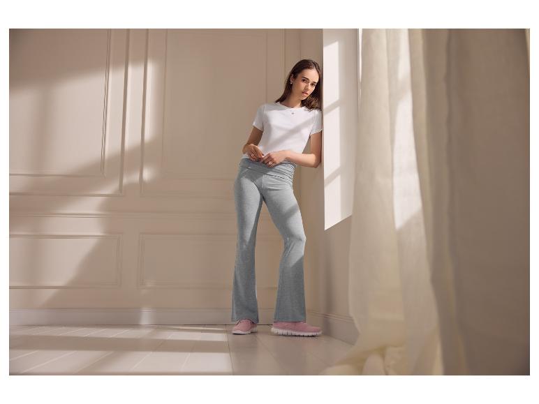 Femme en t-shirt blanc et leggings évasés gris, appuyée contre un mur.