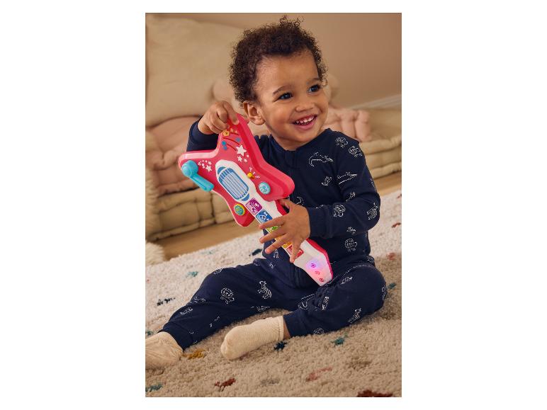 Un bébé souriant en grenouillère bleue joue avec une guitare jouet rose.
