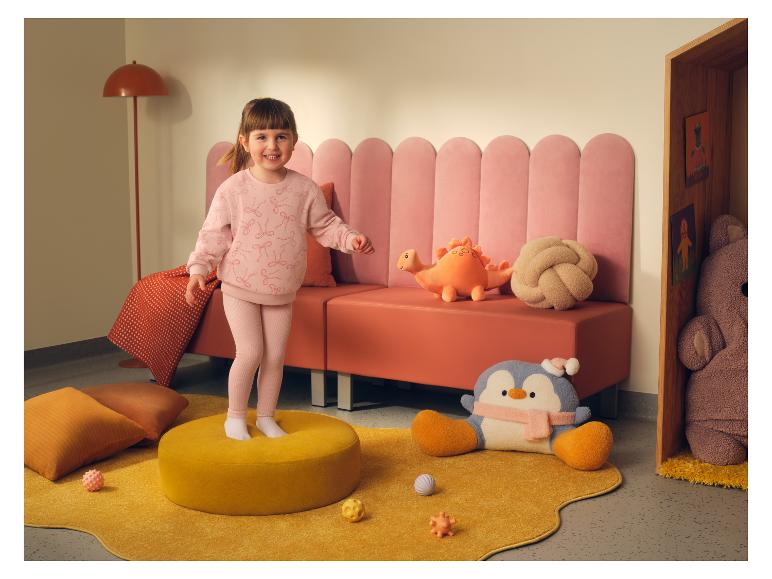 Fillette vêtue d'un ensemble rose LUPILU® avec sweat-shirt et leggings, debout sur un pouf jaune.