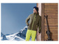 Un homme en tenue de ski avec une veste verte et un pantalon jaune, des skis contre un fond en bois.