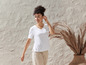 Une femme porte un t-shirt blanc et un pantalon beige devant un mur texturé.