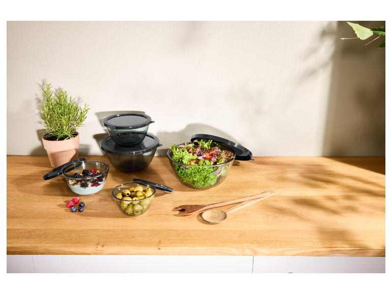 Bols en verre avec couvercles, salade, yaourt aux baies, olives et herbes sur un comptoir en bois.