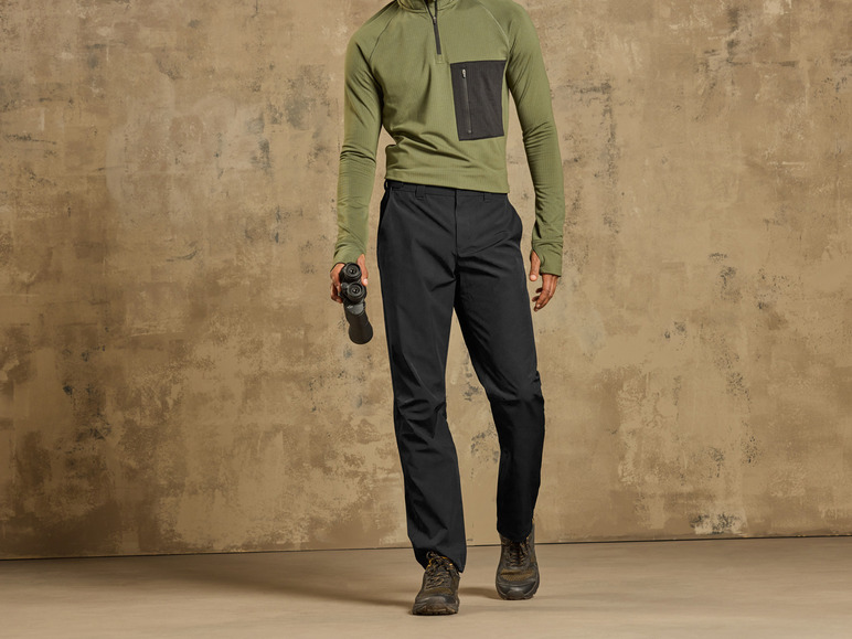 Un homme porte un pantalon noir, un pull vert et tient des jumelles.