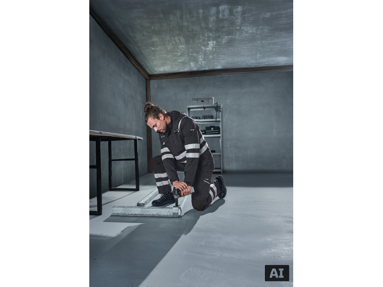 Homme en tenue de travail avec bandes réfléchissantes perçant des profilés métalliques au sol.