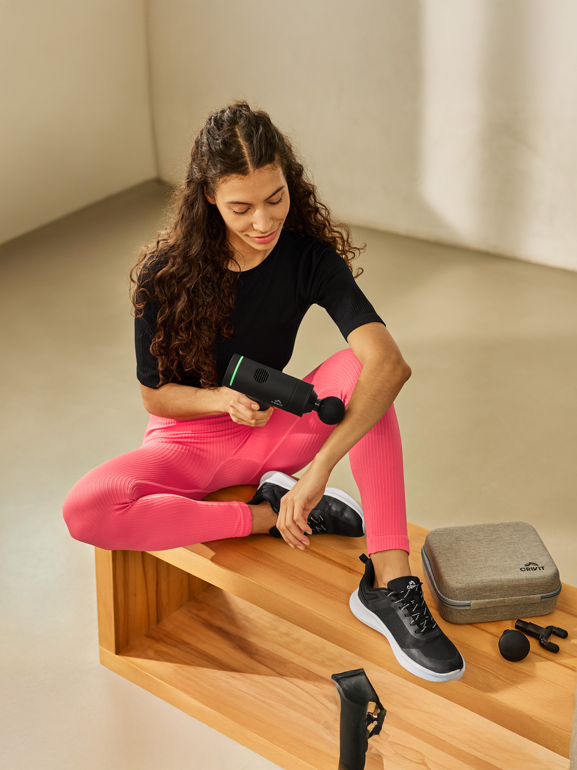 Femme utilise un pistolet de massage, assise sur un banc en bois, à côté d'un étui et d'accessoires.