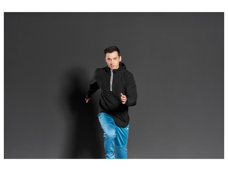 Homme en veste noire et pantalon de survêtement bleu posant sur un fond gris.