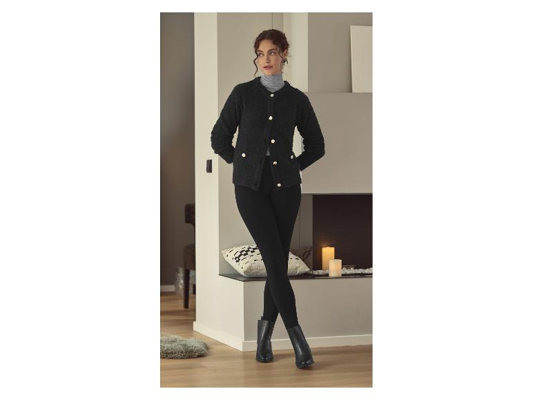 Femme en cardigan noir, leggings noirs et bottes noires, debout près d'une cheminée.