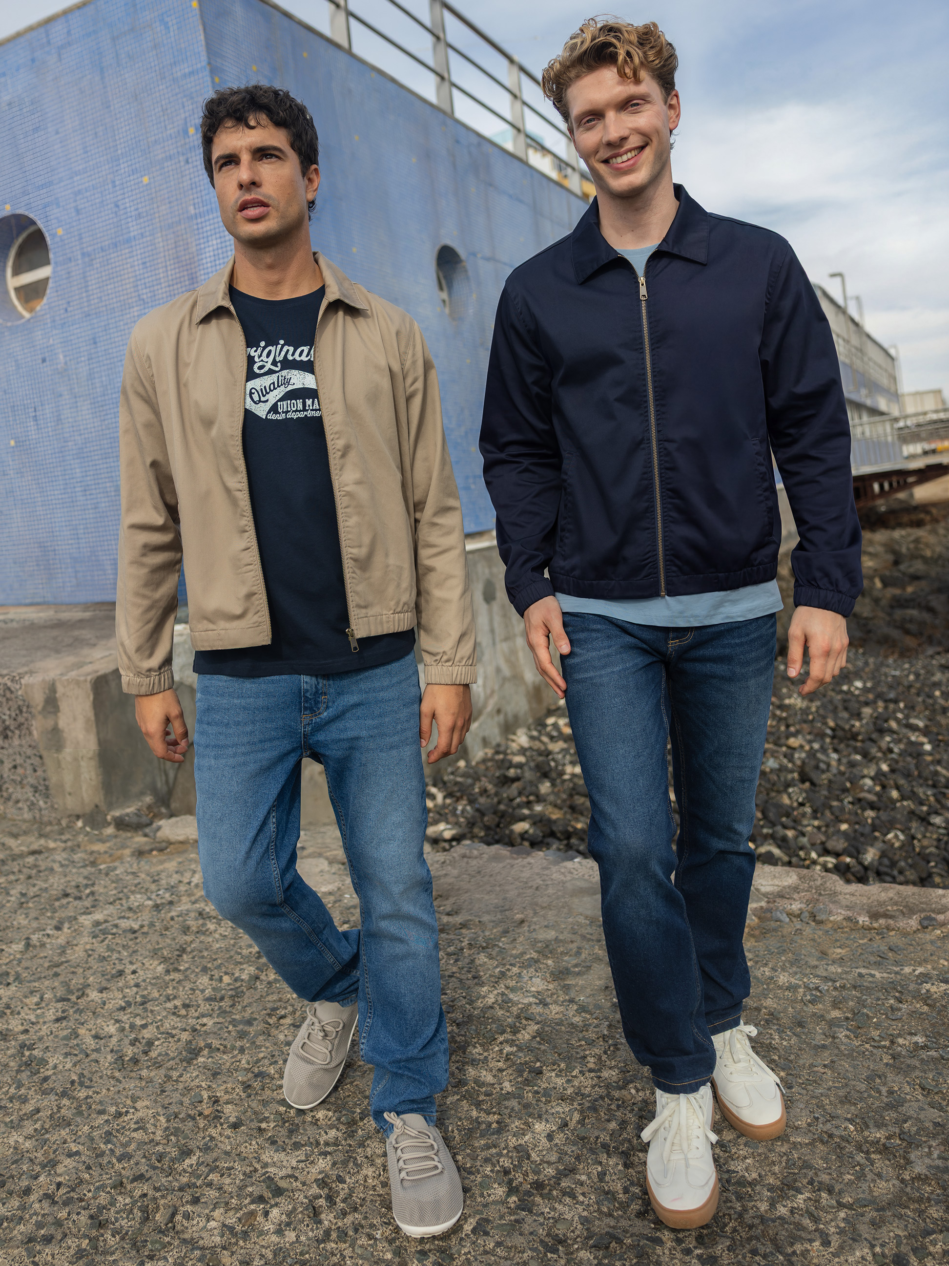 Deux hommes en blousons bomber et jeans, marchant à côté d'un bâtiment bleu.