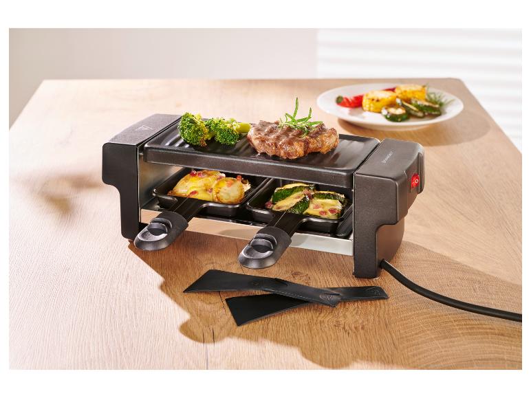 Appareil à raclette avec steak, brocoli et légumes, plus deux spatules sur une table en bois.