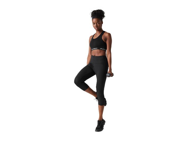 Femme en brassière de sport noire et legging, debout sur une jambe avec un haltère.