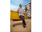 Homme en t-shirt rayé et pantalon foncé appuyé sur un bateau jaune sur la plage.