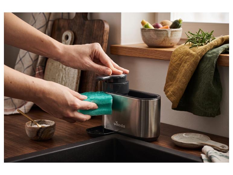Mains pressant du liquide vaisselle sur une éponge verte dans un distributeur en acier inoxydable.