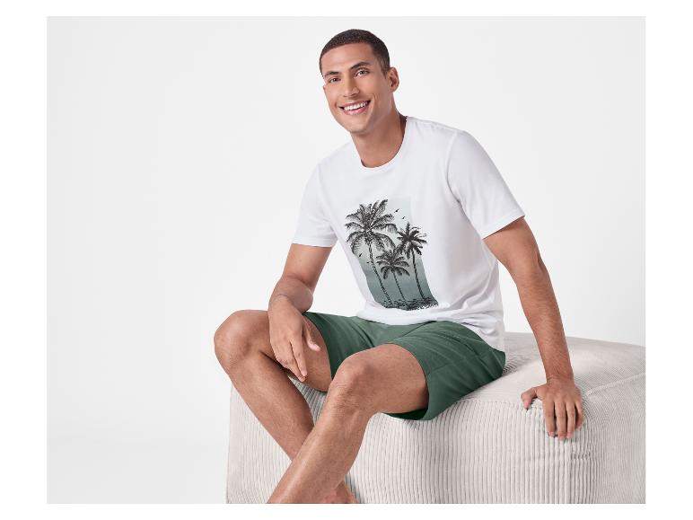 Homme en t-shirt blanc imprimé palmiers et short vert assis sur un pouf.