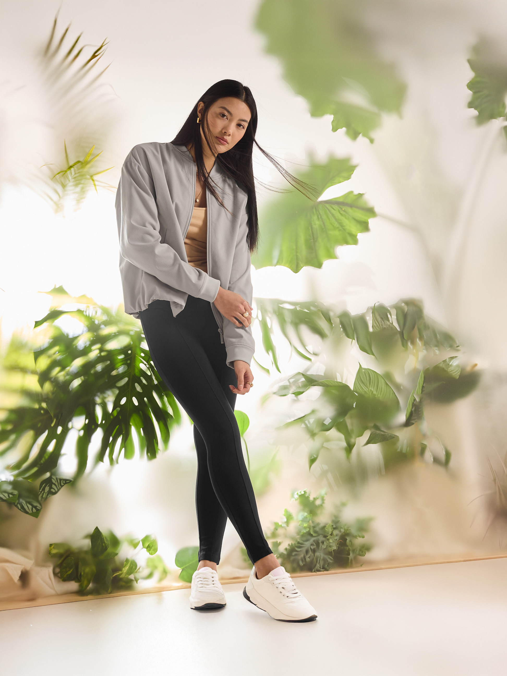 Femme en blouson bomber gris et leggings noirs avec baskets blanches, entourée de verdure.