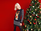 Une femme porte un pull de Noël avec un motif de renne et se tient à côté d'un sapin de Noël.