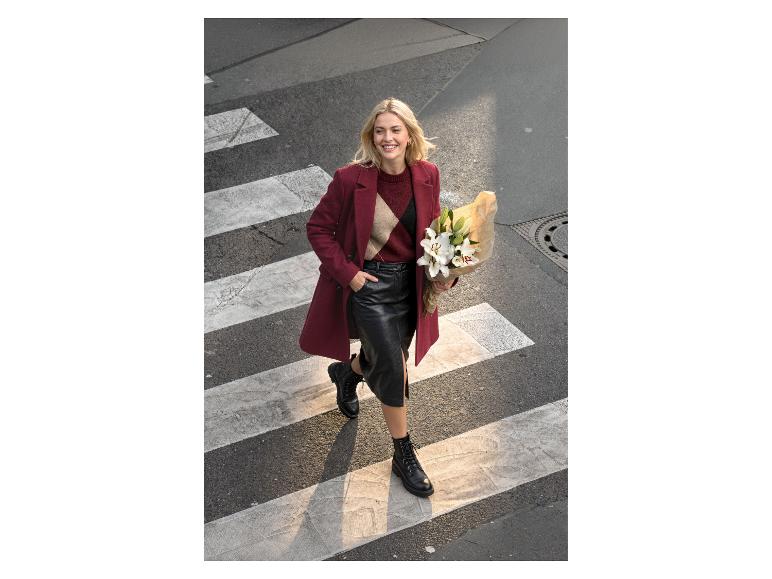Femme en manteau rouge, jupe en cuir noire et bottes traversant un passage piéton avec des fleurs.