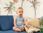 Un bébé dans un body bleu avec un soleil imprimé, assis sur un canapé bleu.