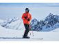 Homme en veste de ski orange, pantalon de ski noir et casque avec masque, debout sur des skis dans un paysage montagneux enneigé.