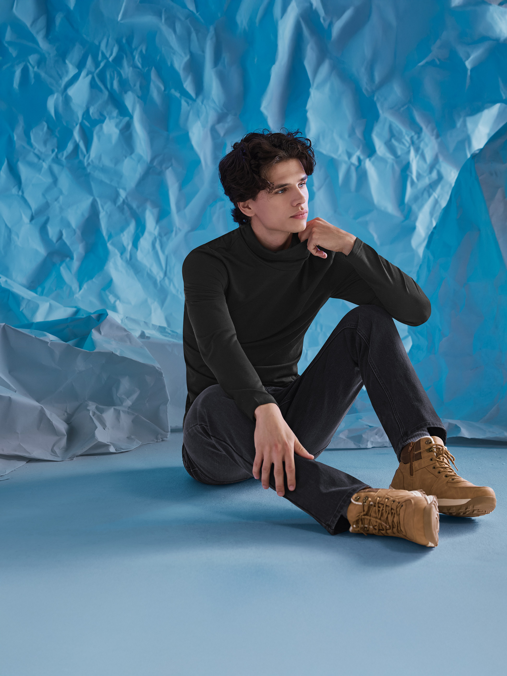 Homme en pull à col roulé noir, jean et bottes marron, assis sur un fond bleu.