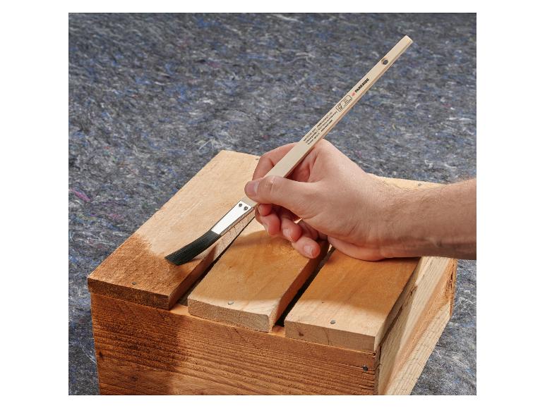 Main appliquant du vernis sur une caisse en bois avec un pinceau, sur un fond gris.