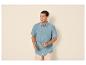 Homme en chemise bleue à manches courtes et short beige, tenant des lunettes de soleil.