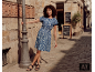 Femme en robe bleue à motifs et sandales noires dans une rue pavée.
