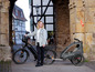 Une femme avec un vélo électrique noir et une remorque Qeridoo dans une ruelle historique.