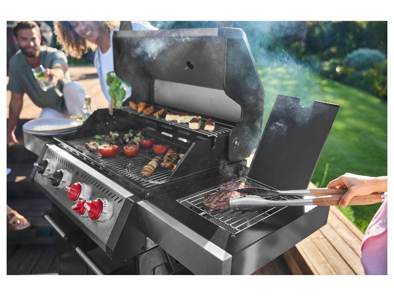 Des amis font griller de la viande et des légumes sur un barbecue à gaz en extérieur.