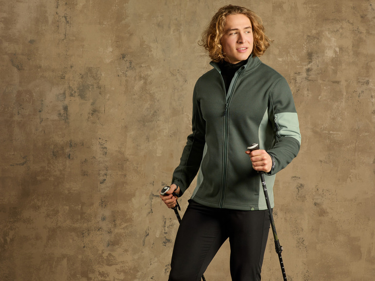 Un homme vêtu d'une veste polaire verte et d'un pantalon noir avec des bâtons de randonnée.