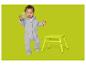 Bébé en grenouillère grise avec jouet vert et tabouret jaune sur fond vert.