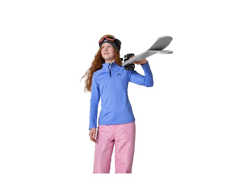 Fille rousse avec lunettes de ski, bonnet, haut bleu et pantalon rose, tenant des skis.