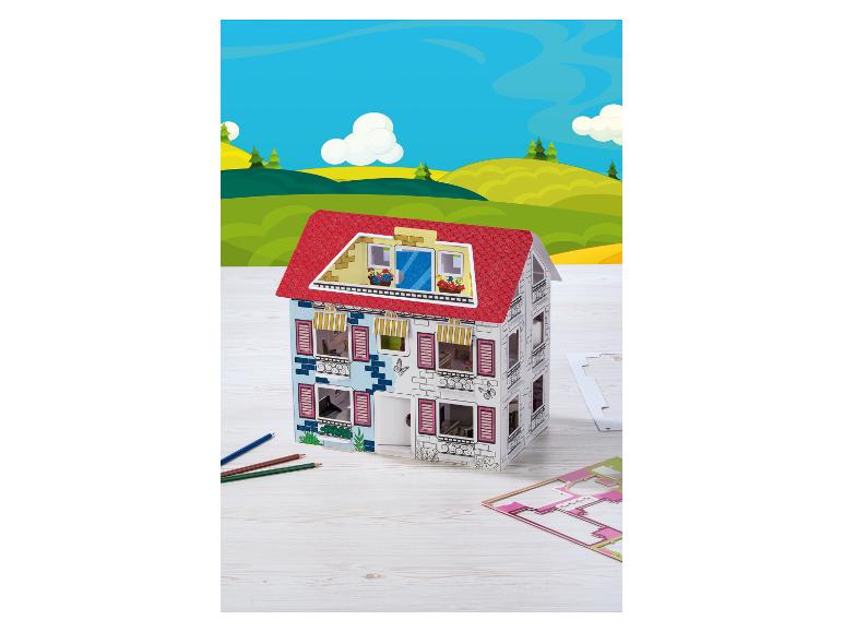 Maison de poupée en carton avec toit rouge, crayons de couleur et matrice de découpe sur une table en bois.