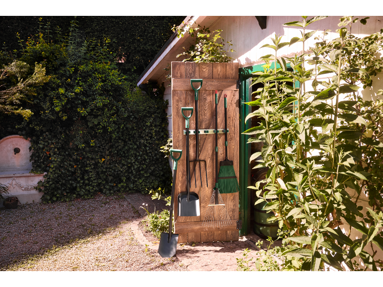 Outils de jardinage suspendus à une porte en bois, avec une pelle et un râteau au premier plan.