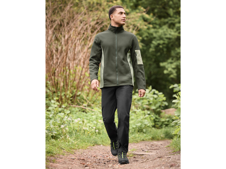 Homme en veste polaire verte et pantalon noir marchant sur un sentier.