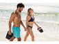 Couple marchant sur la plage, homme en short de bain tenant des claquettes, femme en bikini tenant des claquettes.