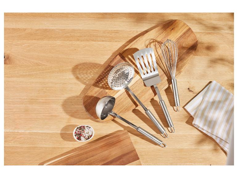 Ustensiles de cuisine en acier inoxydable, dont une spatule, une louche et un fouet, sur une planche à découper en bois.