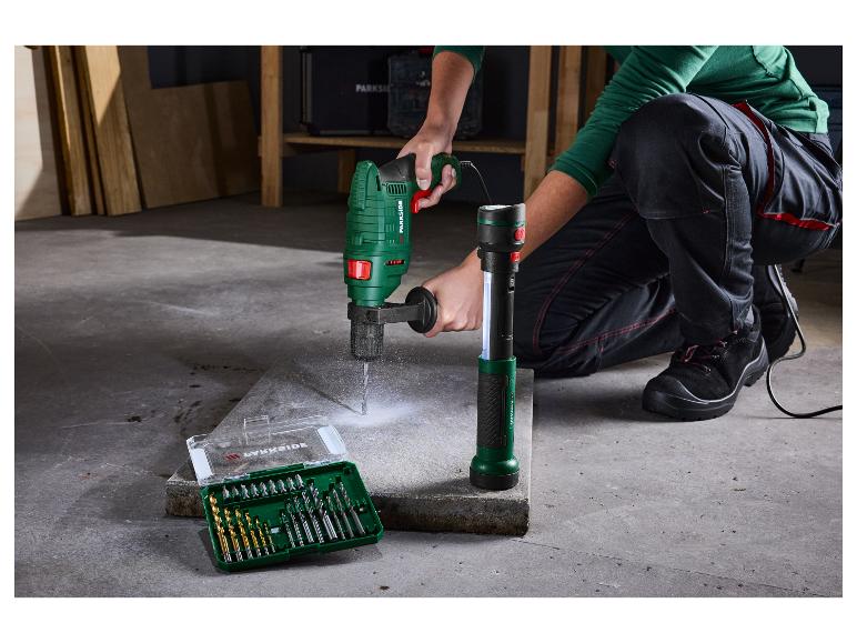 Une personne utilise une perceuse verte sur une dalle de béton, avec un jeu de forets et une lampe de poche à côté.