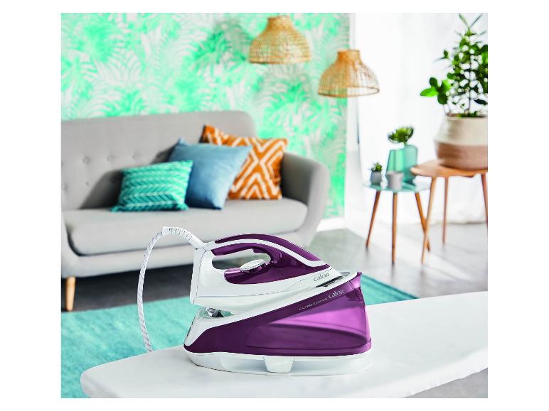 Fer à vapeur violet sur une planche à repasser dans un salon avec plantes et canapé.