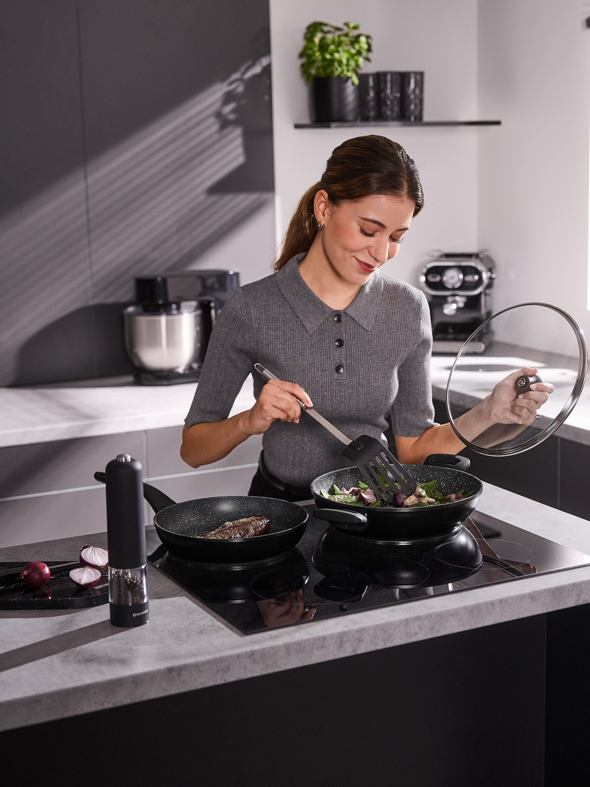 Femme cuisinant avec deux poêles sur une plaque à induction, tenant un couvercle et une spatule.